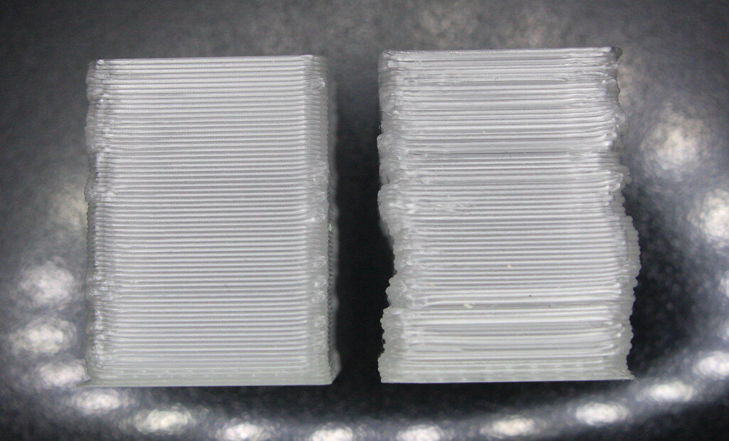 Two translucent printed blocks side by side: the left with even layers, the right with wavy, shifted walls from a pulley problem