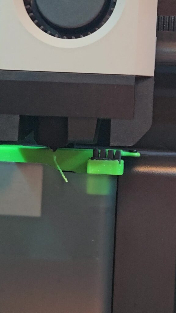 3D printer nozzle with a thin green filament string dangling from the tip, a typical sign of stringing and oozing
