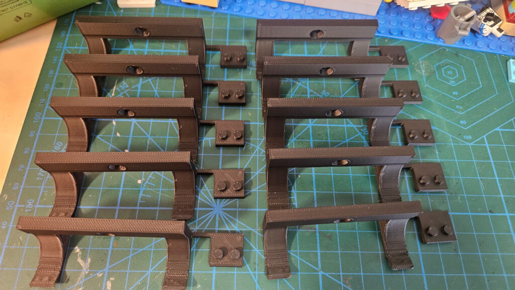 Ten dark printed brackets with curved arms and small pegged tabs laid out on a green cutting mat