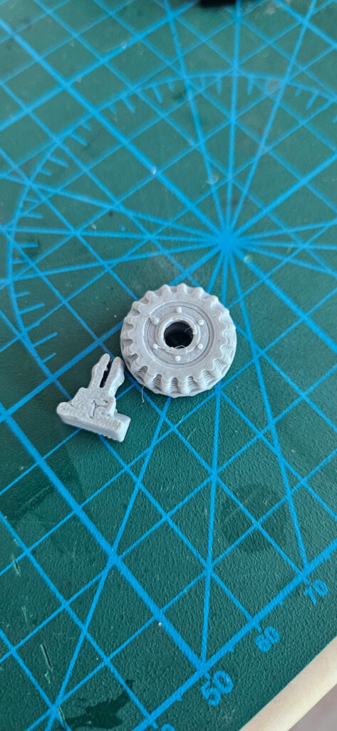 Small grey printed clip with two split arms next to a printed toothed wheel on a cutting mat