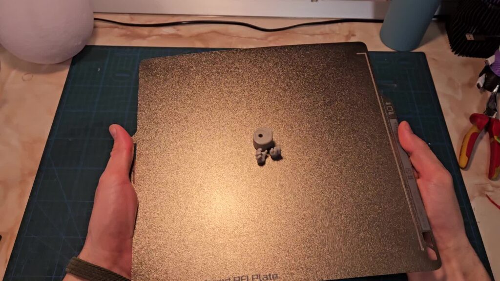 Hands holding a textured PEI build plate by its edges, with small grey printed parts in the middle