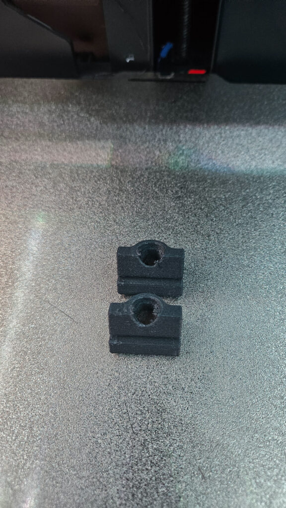 Two black printed brackets with round holes sitting on a textured print bed, layer lines visible on their sides