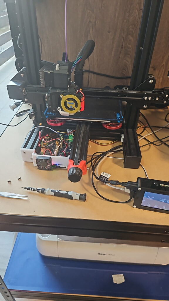 Biqu B1 bed slinger on a desk: the bed rides on a Y rail with a front belt tensioner knob, electronics box open beside it