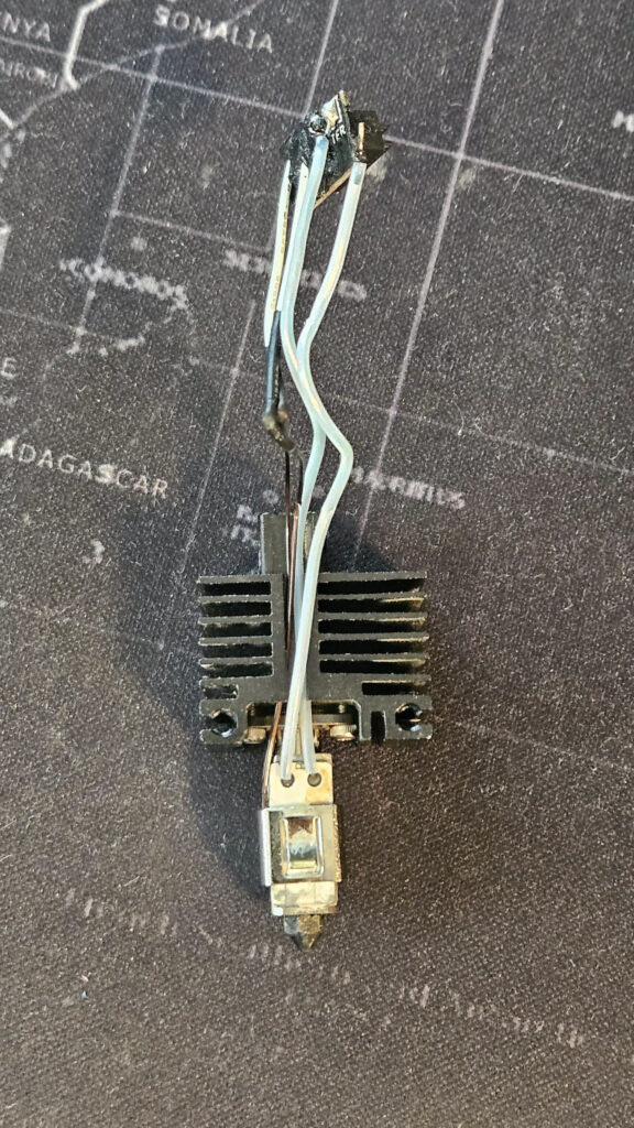 Removed 3D printer hotend with heatsink, heater block, nozzle and exposed heater and thermistor wiring laid on a dark mat