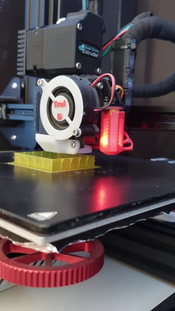Close-up of a 3D printer's blower fan and printed duct cooling a yellow part being printed on a Creality bed