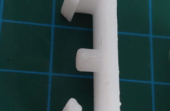 White 3D-printed snap-fit clip with hooked ends and a small peg, lying on a green cutting mat