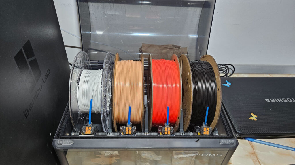 Bambu Lab AMS beside a P1S, loaded with white, beige, red and black filament on plastic and cardboard spools