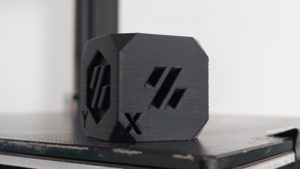 Black printed test cube with a chamfered edge, an X label and recessed logo, showing even layer lines and crisp edges