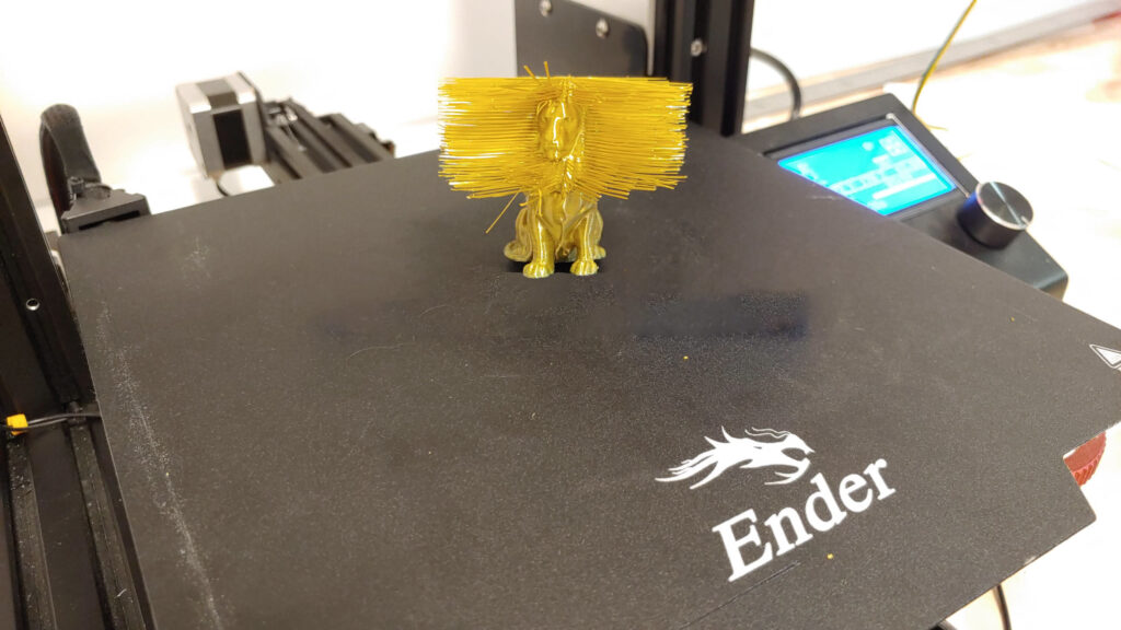 Small yellow lion figure on a Creality Ender print bed, almost hidden behind a dense curtain of fine plastic strings